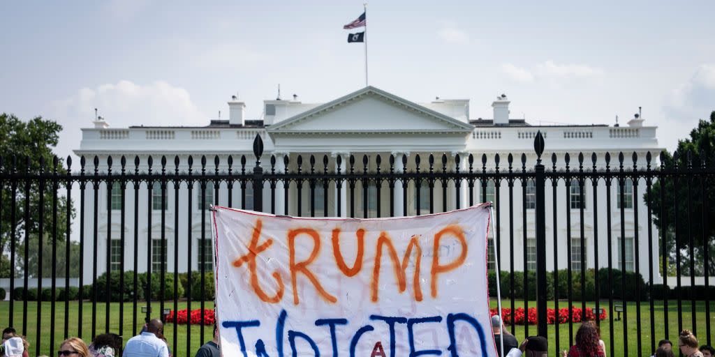 a group of protestors standing outside the gate of the white house, one of whom holds up a sign that reads trump indicted