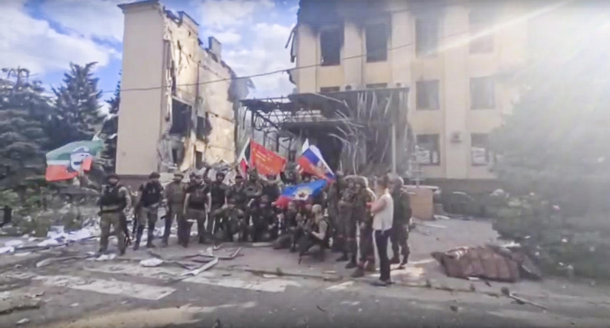 This photo taken from video provided by Ramzan Kadyrov's Official Telegram channel released on Saturday, July 2, 2022, shows Russian troops including soldiers of Chechen regiment waving Russian and Chechen republic national flags as they pose for a photo in front of a destroyed building in Lysychansk, Ukraine.