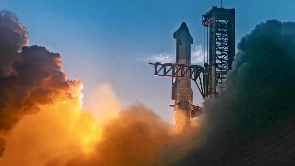 The top of a giant steel rocket just after launch next to the black launch tower. Smoke glowed with orange waves below, filling the frame on both sides.