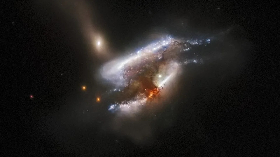  A telescope image of three galaxies swirling around one another as they collide, with a bright orange burst of radiation at the center 