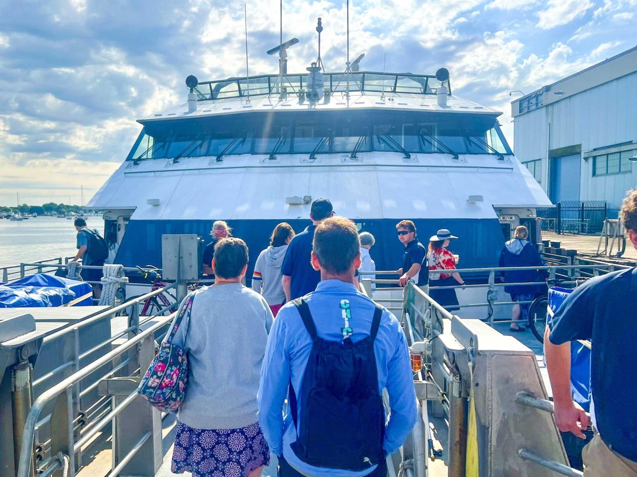 Several people on a dock lined up to board a large white and blue ferry. A man in the foreground wears a blue jacket and black backpack.