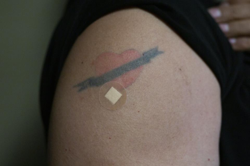 Un bras portant un tatouage de cœur et un petit pansement rond sur un point d’injection