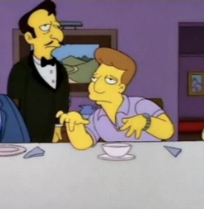 A waiter in a suit stands at attention, while Freddy sits at a table making a face and a funny hand gesture