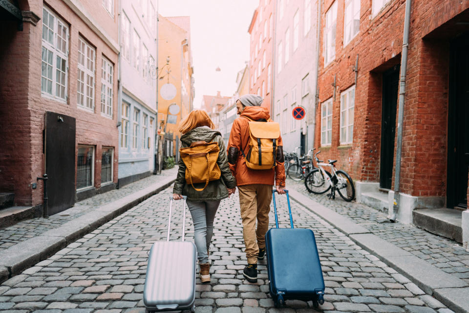 Two travelers with yellow backpacks and rolling suitcases walk down a cobblestone street in a European city