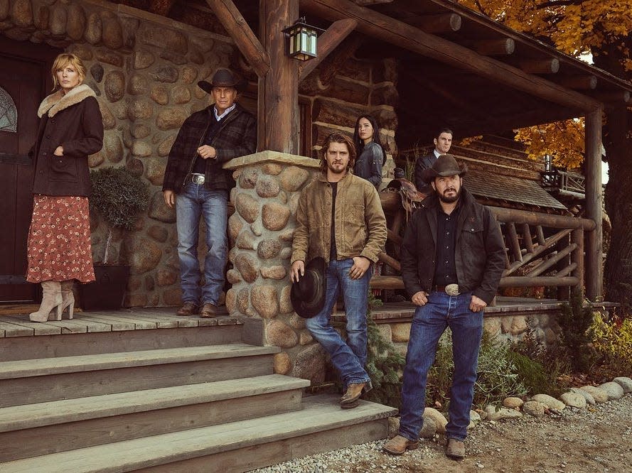 The cast of "Yellowstone" posing on the porch of a simple-looking house with stone accents and multiple lanterns.