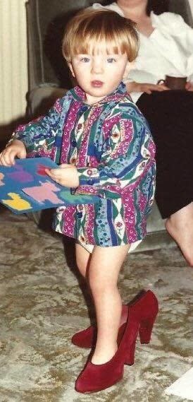 A toddler wearing a woman's high-heeled red shoes and a colorful top