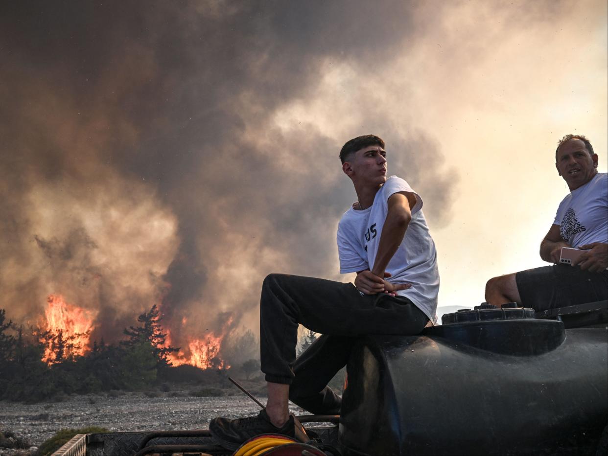 Men sit on the back of their pick-up truck as wildfires rage in the background close to the village of Vati on the Greek island of Rhodes on 25 July (AFP)