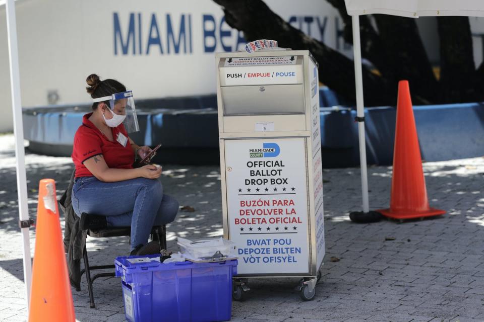 A woman with a mask on sits near an election drop box.