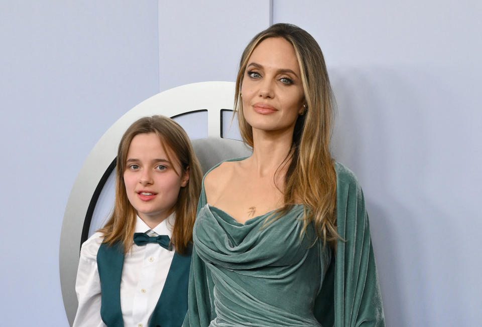 Angelina Jolie and her daughter Vivienne pose together on the red carpet. Angelina is wearing an elegant draped dress, and Vivienne is dressed in a vest and bow tie