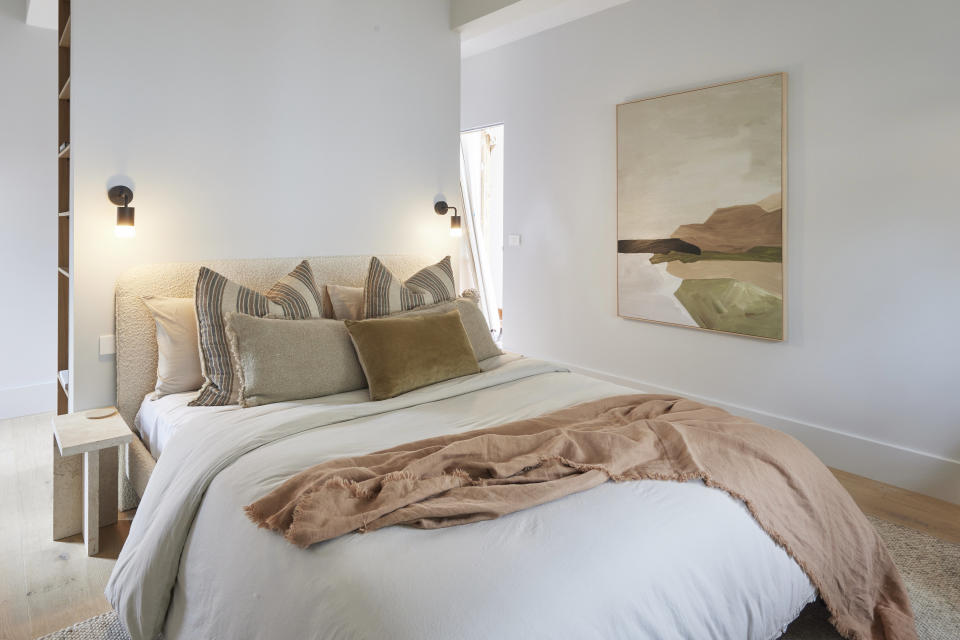 A close up of the bed with a light brown throw, and a painting on the far wall. 
