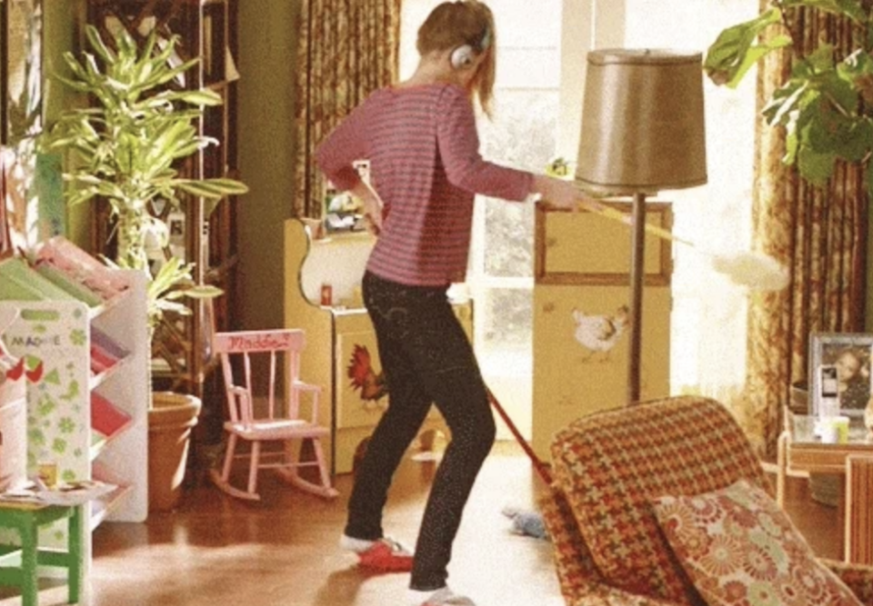 Woman wearing headphones dusting, mopping and dancing in a living room filled with plants, furniture, and children's toys