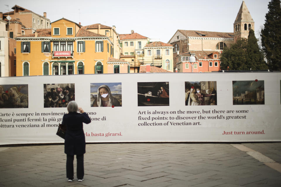 A tourist takes a photograph of a board promoting art that has been modified by adding masks, in Venice, Saturday, Feb. 29, 2020. A U.S. government advisory urging Americans to reconsider travel to Italy due to the spread of a new virus is the "final blow" to the nation's tourism industry, the head of Italy's hotel federation said Saturday. Venice, which was nearing recovery in the Carnival season following a tourist lull after record flooding in November, saw bookings drop immediately after regional officials canceled the final two days of celebrations this week, unprecedented in modern times. (AP Photo/Francisco Seco)