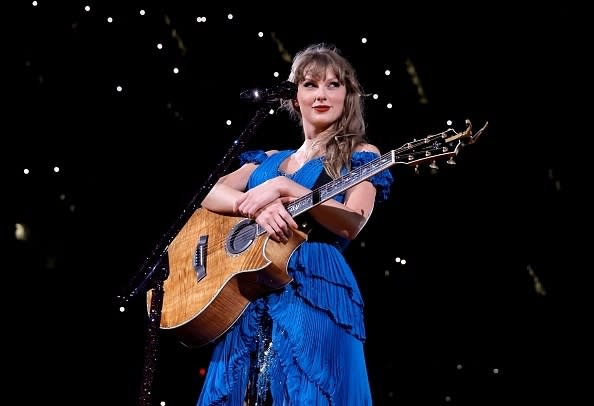 Taylor Swift performs on stage with a guitar, wearing a fringed blue dress