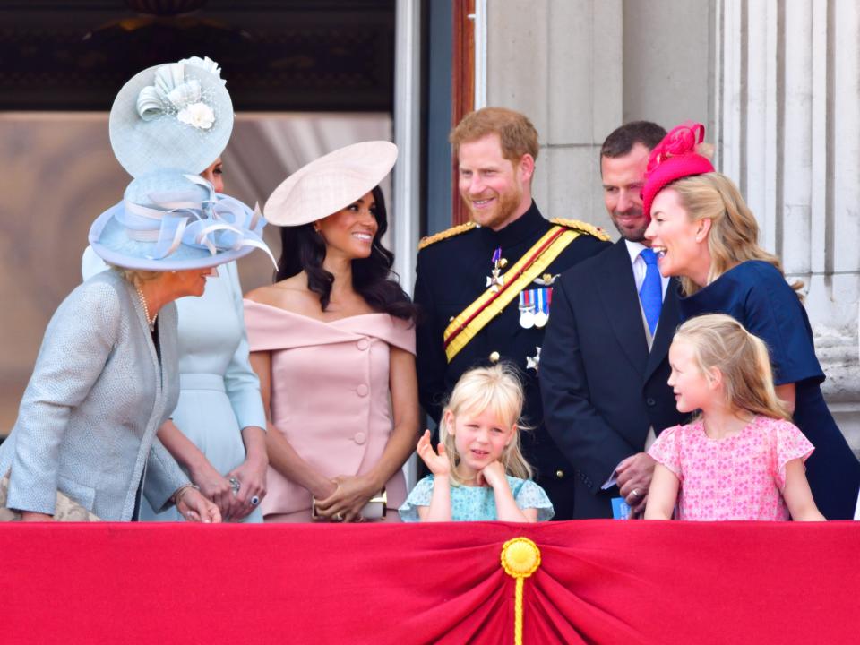 Meghan in a pink dress and fascinator and Harry in a military uniform both smiling towards each other surrounded by family looking and speaking to them on a balcony.