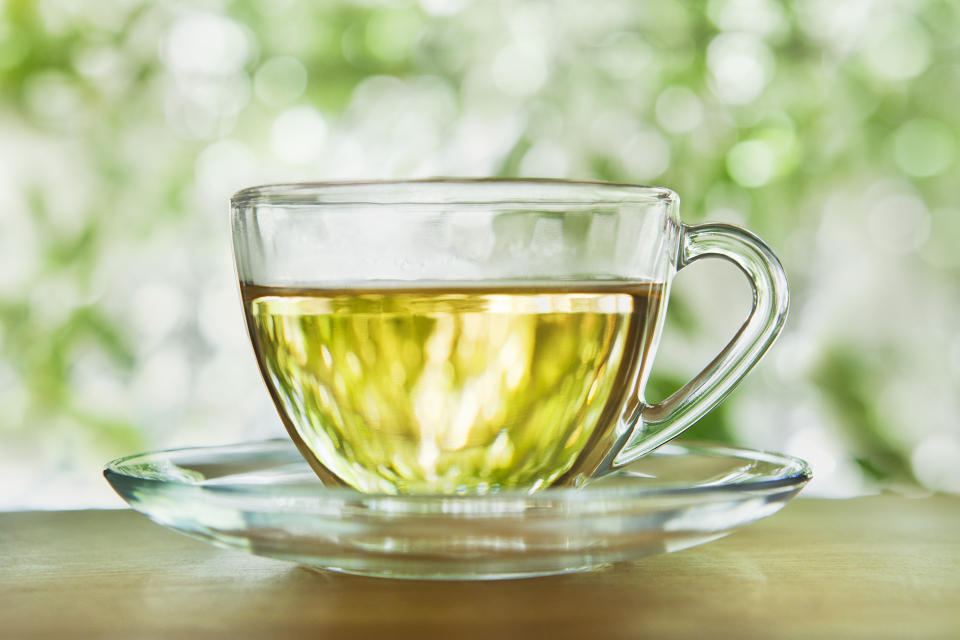 A glass cup and saucer with herbal tea against blurred foliage outdoors.