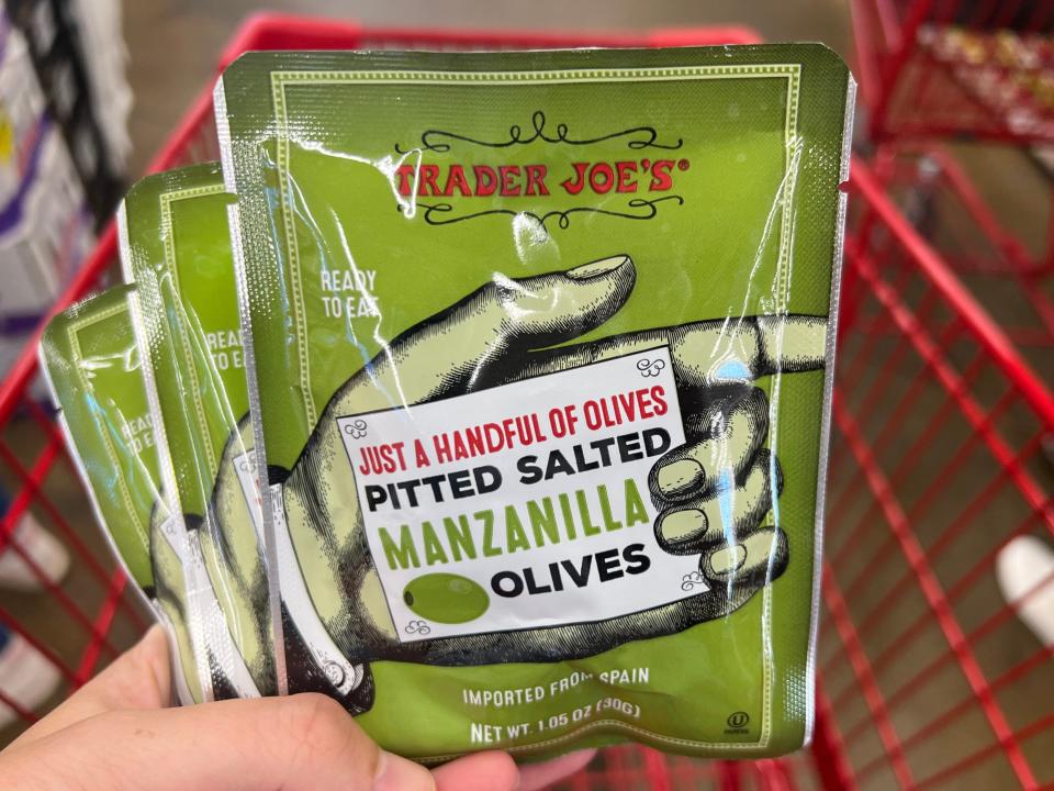 The writer holds three green packages of olives with an illustration of a hand and Trader Joe's logo on it