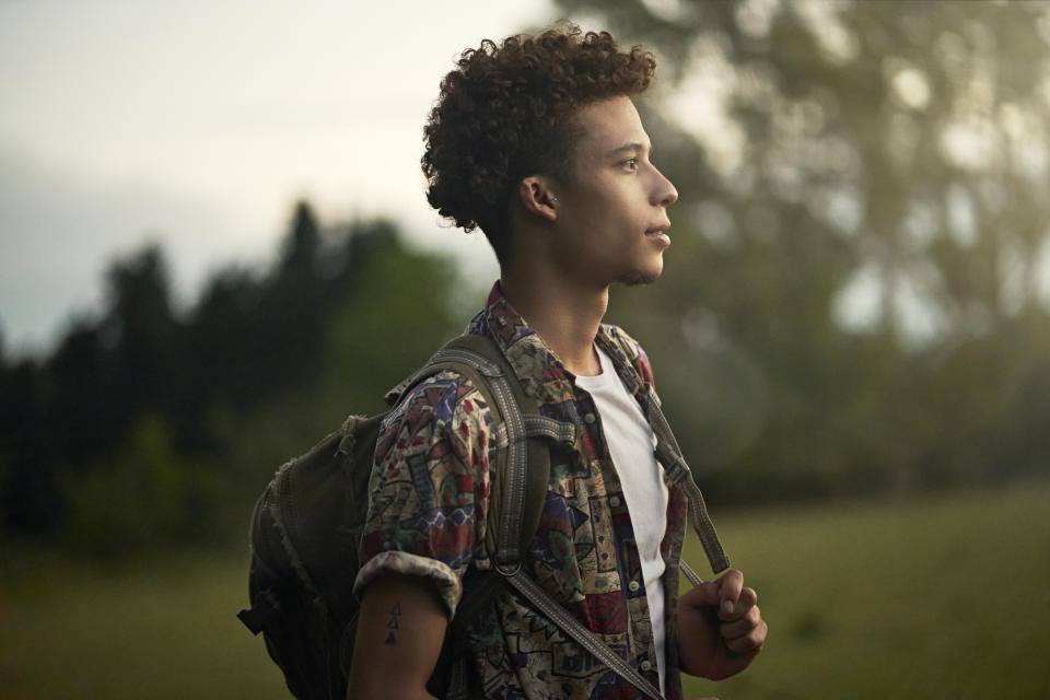 Waist-up profile view of contemplative man with curly brown hair wearing casual clothing, backpack, and looking away from camera at dusk.