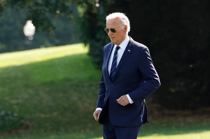 Joe Biden, wearing a suit and tie with sunglasses, walking outside on grass with greenery in the background