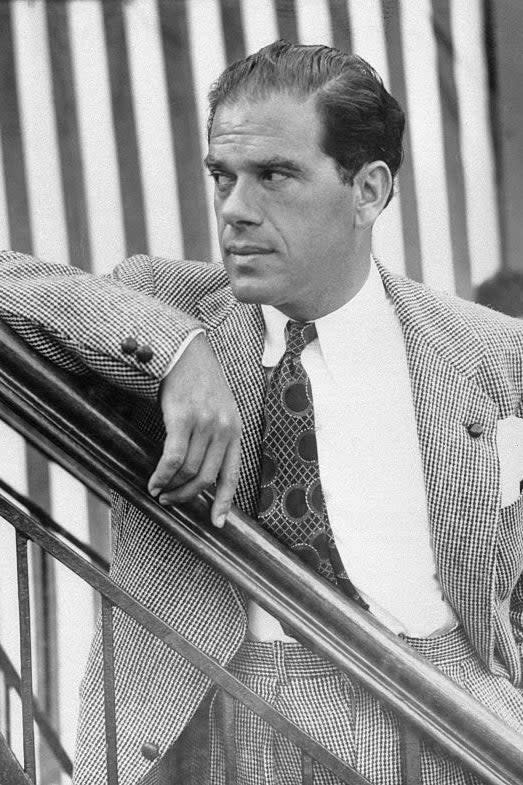 Frank Capra in patterned suit with tie leaning on railing, looking thoughtful. Vintage film still