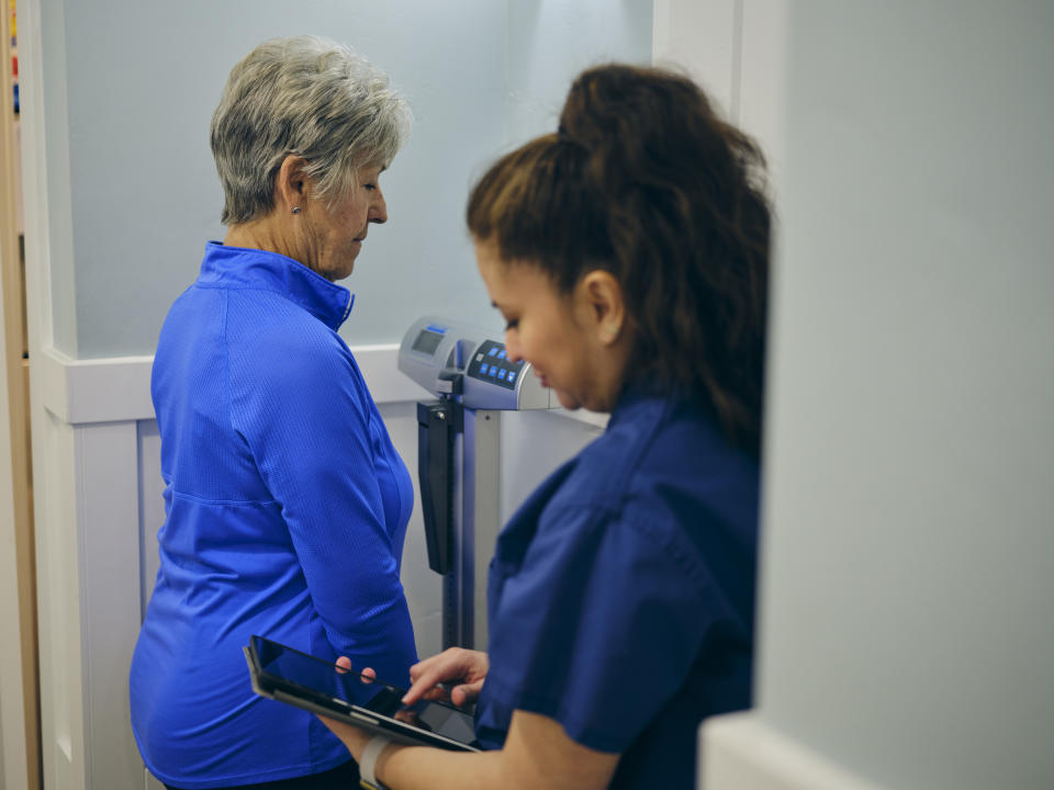 A nurse in a doctor’s office, wearing a senior female patient on a scale.