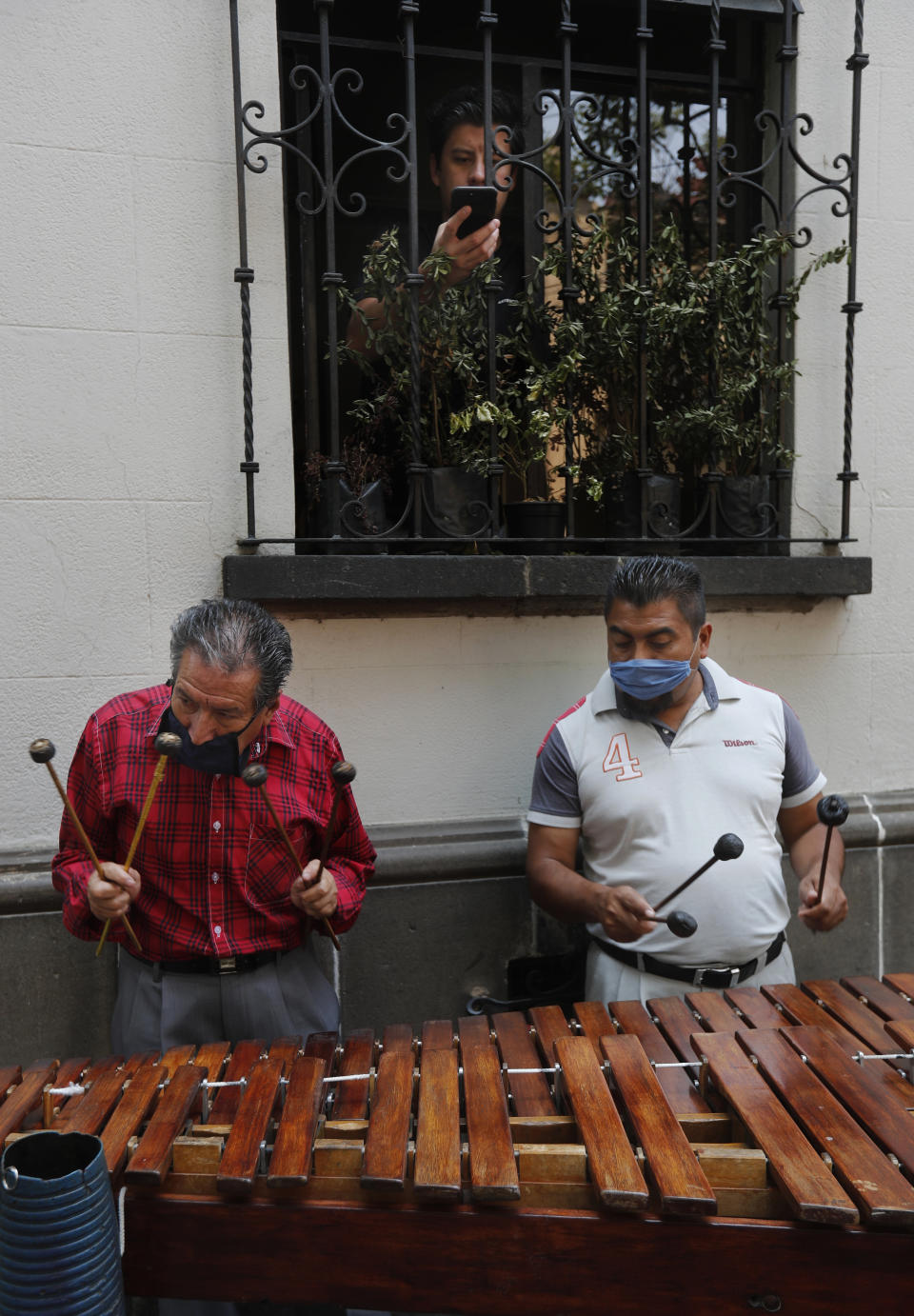Marimba musicians wearing protective face masks perform outside an apartment building as a resident records their performance from inside his apartment, in Mexico City, Thursday, April 16, 2020. The wandering musicians, who usually perform at restaurants, have taken to performing in neighborhoods in hopes that the residents in voluntary lockdown will tip them, which many do. (AP Photo/Marco Ugarte)