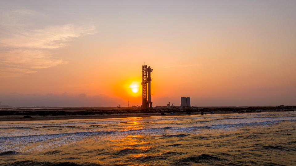 view of a giant rocket on the launch pad with the ocean in the foreground and the setting sun in the background