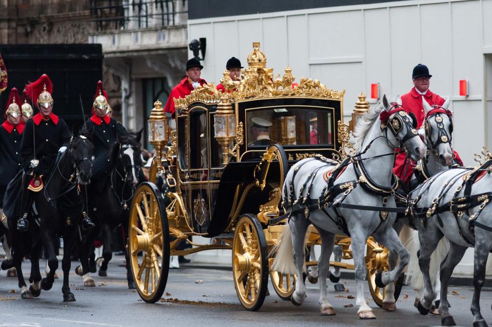 a black and gold carriage is pulled by white hoses, with several men in red uniforms directing it, followed by men in black uniforms on black horses