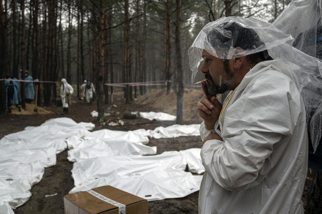 Expert smokes under the rain during the exhumation of bodies in the recently retaken area of Izium, Ukraine, Friday, Sept. 16, 2022. Ukrainian authorities discovered a mass burial site near the recaptured city of Izium that contained hundreds of graves. It was not clear who was buried in many of the plots or how all of them died, though witnesses and a Ukrainian investigator said some were shot and others were killed by artillery fire, mines or airstrikes. (AP Photo/Evgeniy Maloletka)