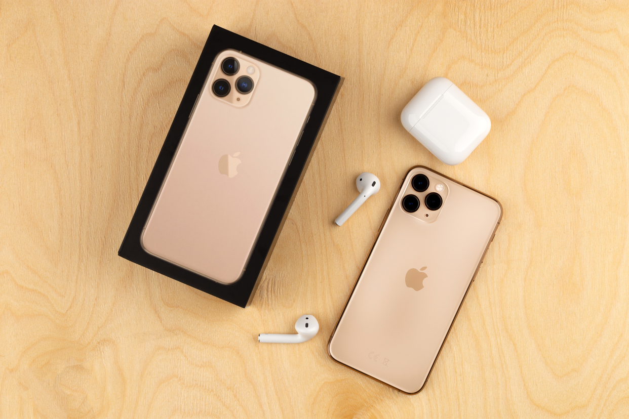 Two rose gold Apple iPhone 11 Pro smartphones with a pair of Airpods earphones on the table, one iPhone still in an opened Apple iPhone box
