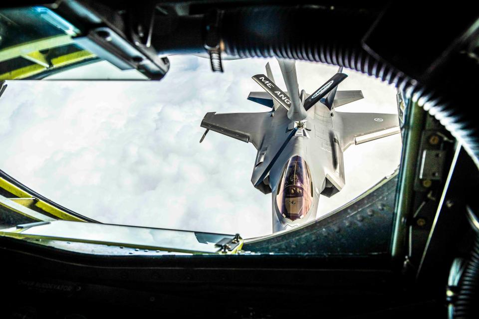 An F-35 fighter jet flies above a white, cloudy background. In the foreground, a ship's refueling section frames the photo.