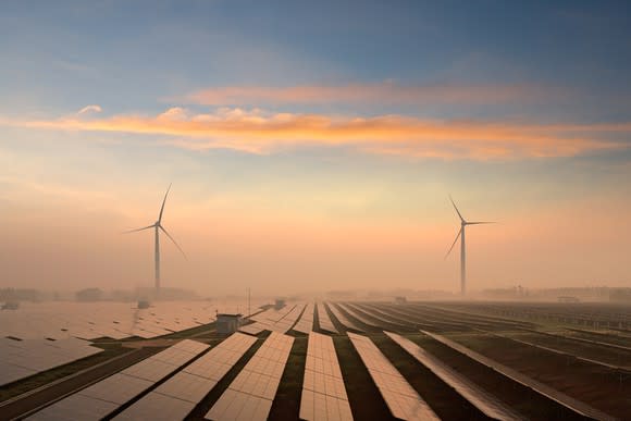 Solar panels spread out over the horizon with wind turbines in the distance.