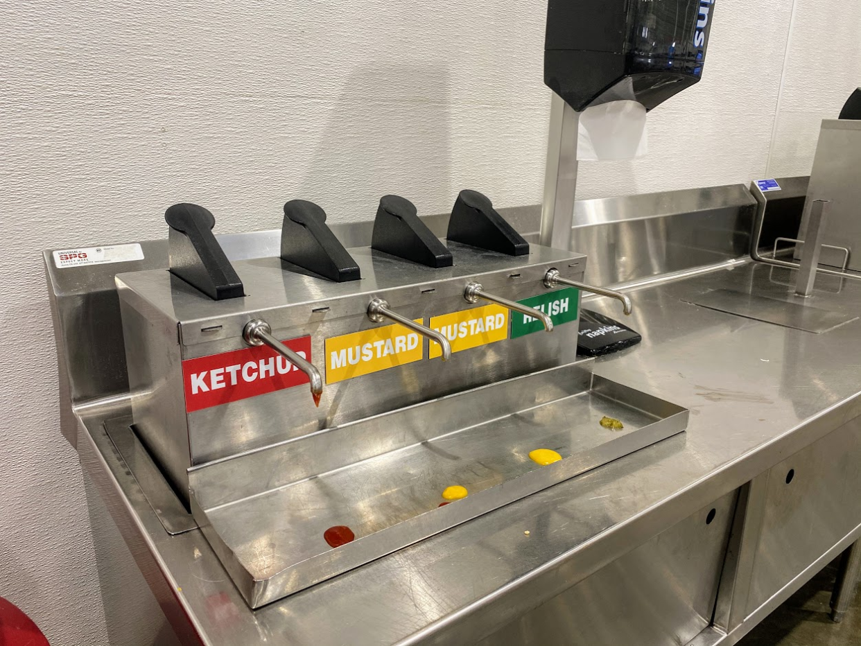 Condiments area for the food court at Costco, four pumps for ketchup, two mustards ones, and relish, and napkins, clean stainless steel table, white wall