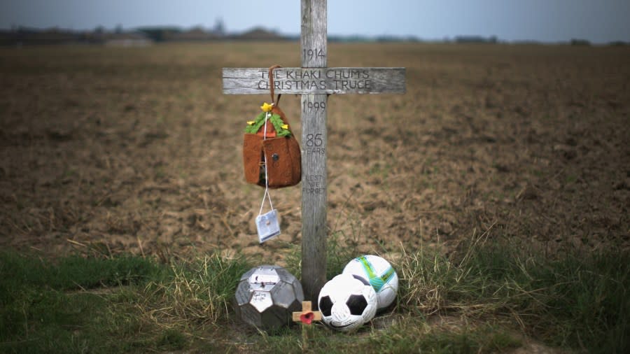 A simple wooden cross stands before an open field. Three soccer balls lay at its base, along with another cross with a red poppy. The cross reads "The Khaki Chums Christmas Truce, 1914, 1999, 85 years, lest we forget."