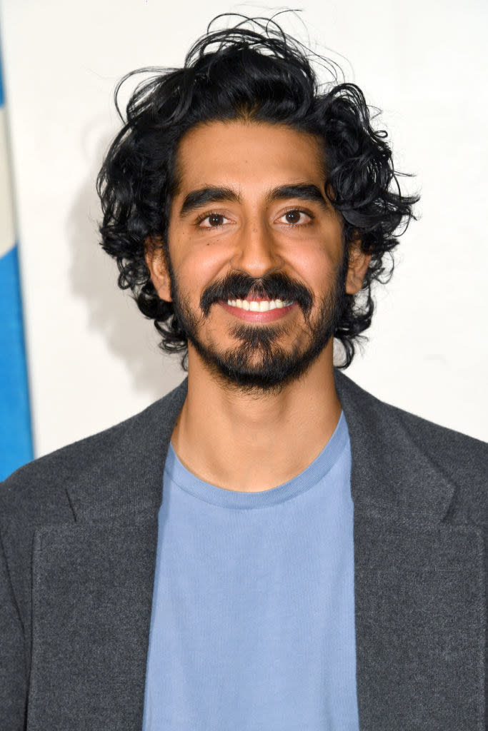 Dev Patel on the red carpet, smiling with long hair and a beard