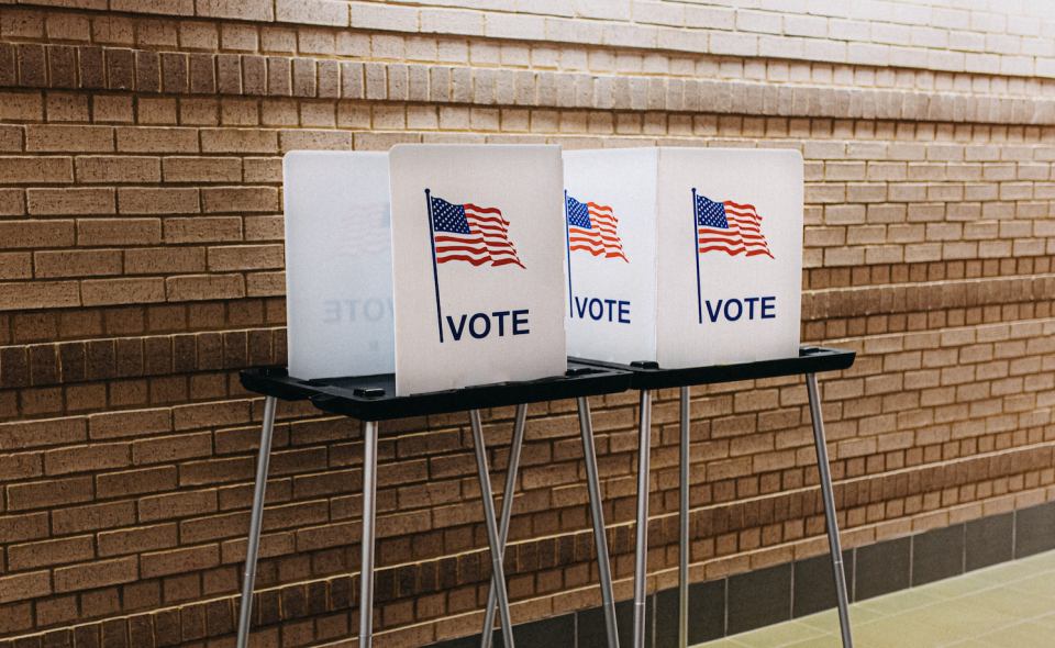 Three voting booths with privacy screens and American flags against a brick wall, indicating a polling place