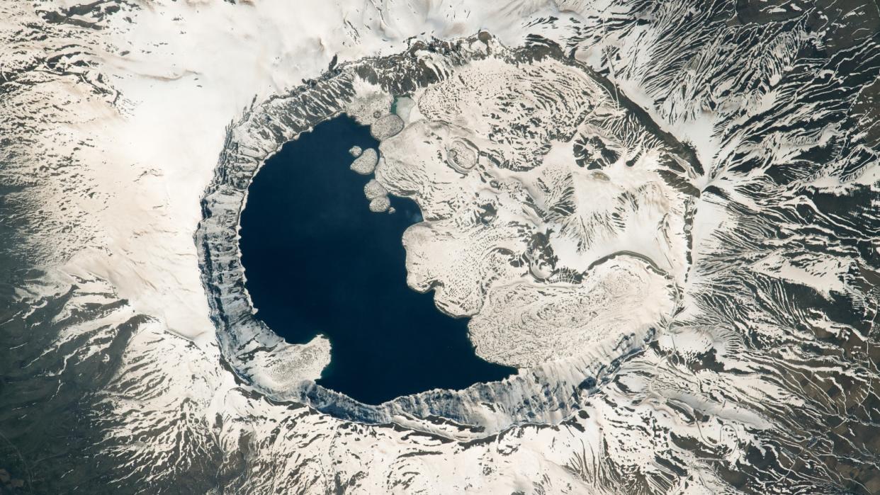  Satellite photo of a sow covered volcano with a larger crater filled with a lake and lava flows that make it look like a yin-yang symbol. 