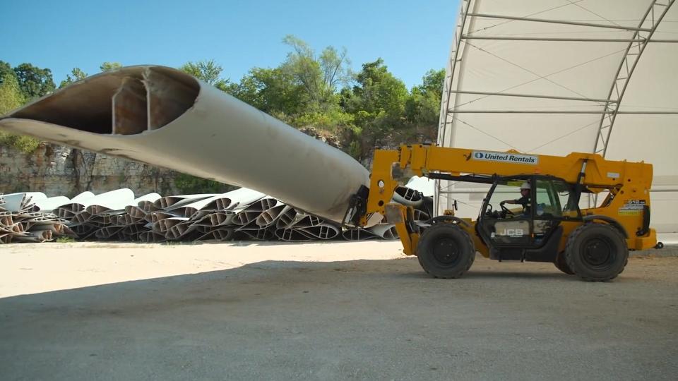 Side view of a worker driving a yellow construction vehicle which is carrying a large wind turbine blade.