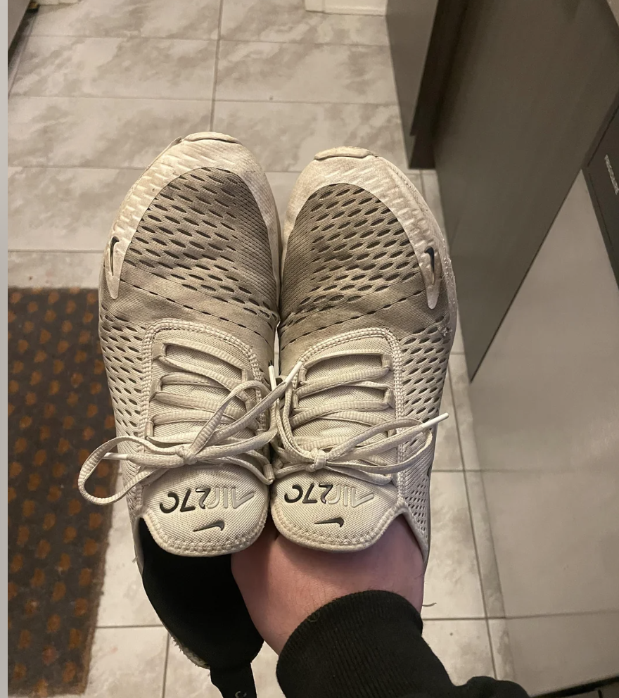 Hand holding a pair of worn athletic shoes with visible signs of use on the uppers and soles. The shoes are placed toe-to-toe. No text or celebrities present