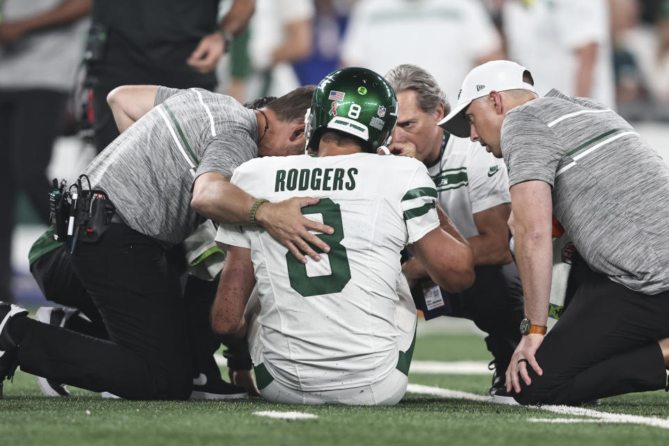 Aaron Rodgers, seated on turf, is evaluated by medical staff.