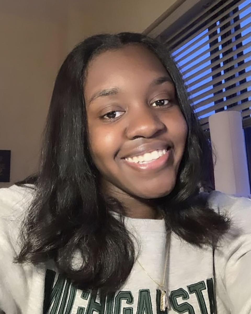 A vertical head-and-shoulders selfie of a young, smiling woman with shoulder-length hair, wearing Michigan State sweatshirt.