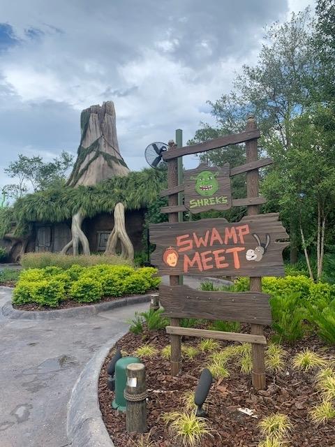 Image shows a wooden sign reading "Shrek's Swamp Meet" with painted characters from Shrek. In the background, there is a cottage with a tree-like appearance