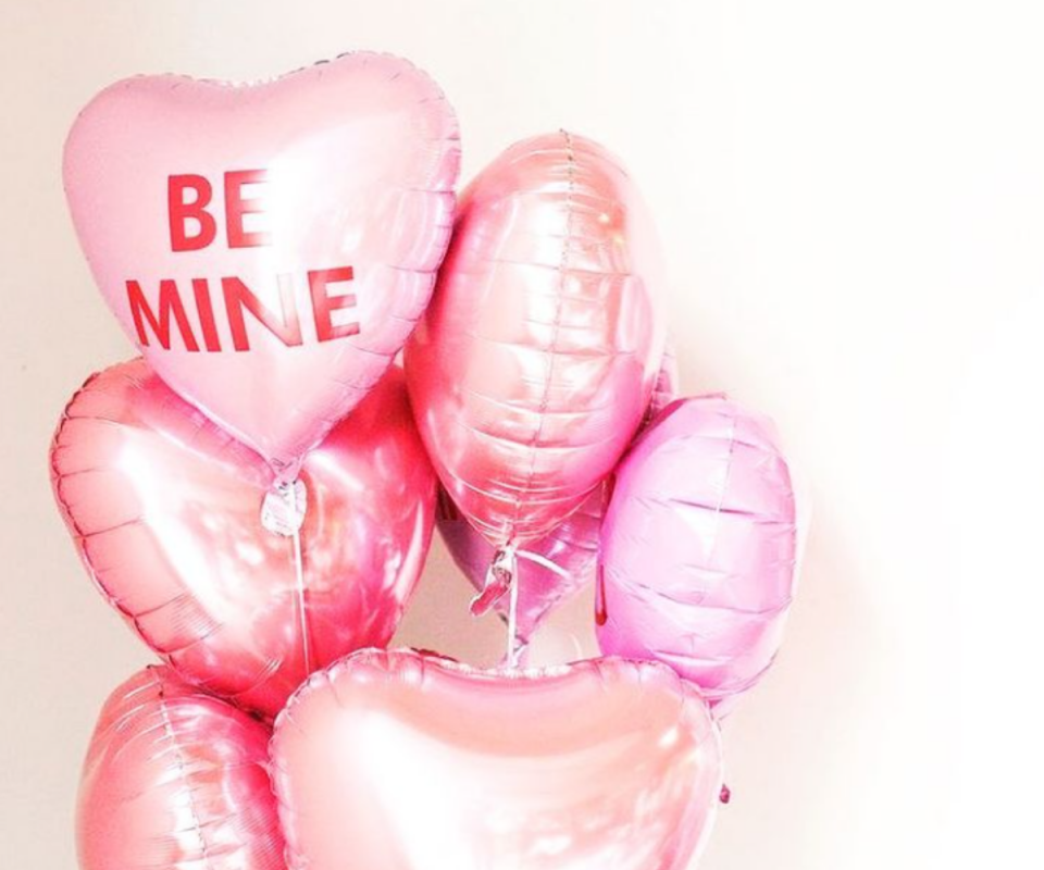 A collection of shiny pink love heart balloons against a white wall with the words, Be Mine in red capitals on the front pink balloon.