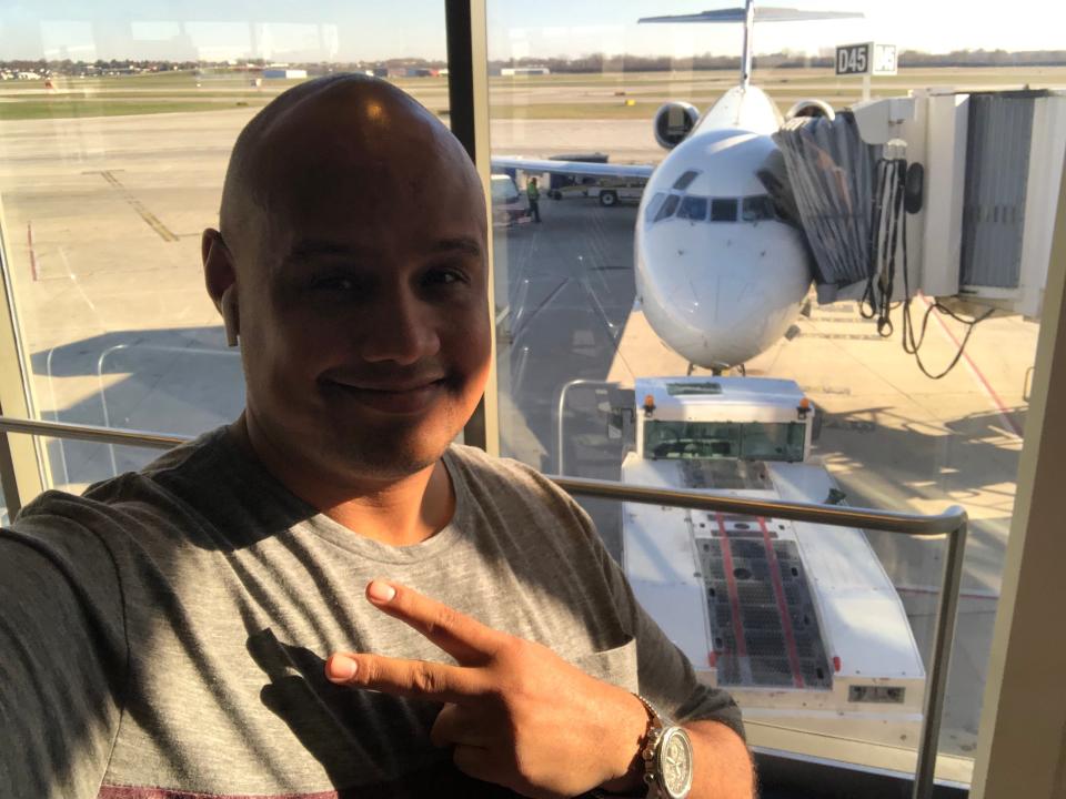 A man taking a selfie and giving a peace sign next to a view of a plane out the window at an airport.