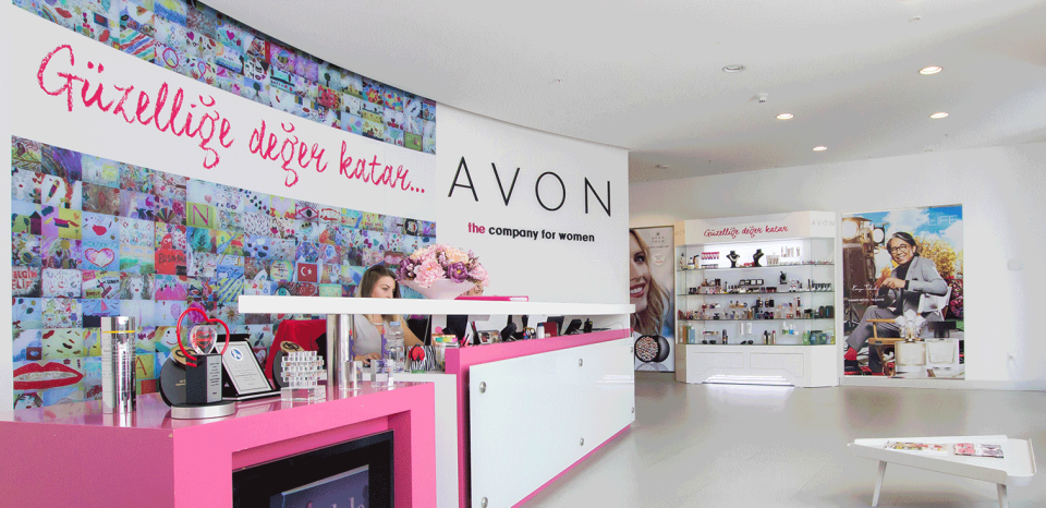 Office lobby with Avon logo on wall, with white and pink desk and various pictures and products.