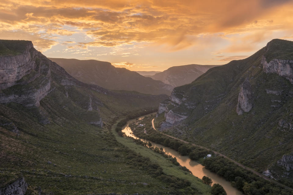 a river snakes between large hills with a pale orange hued partly cloudy sky.