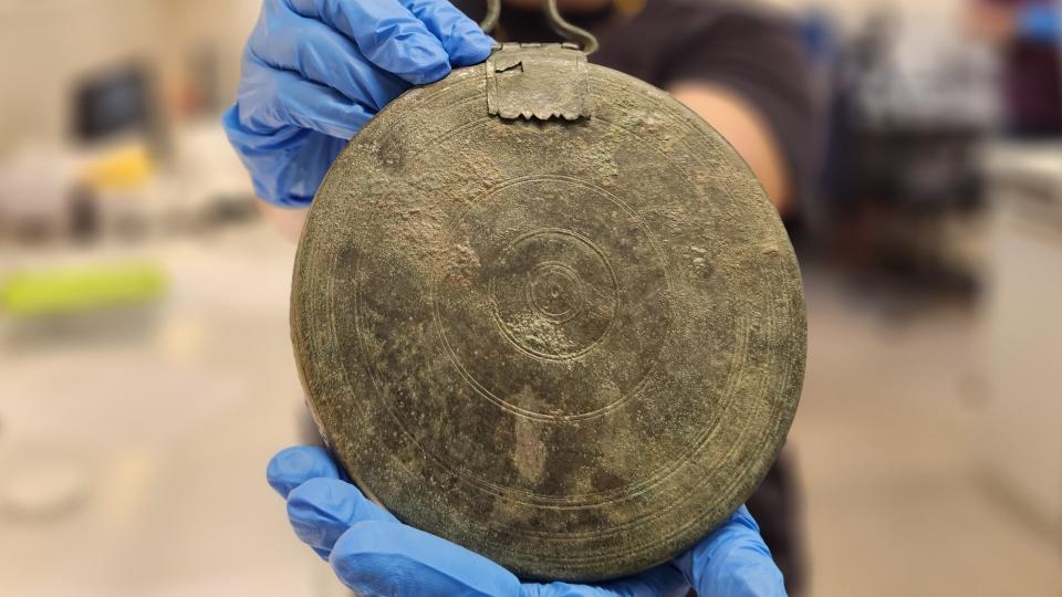 Gloved hands hold up the bronze folding mirror, showing its engravings.