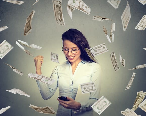 A woman checking her smartphone and pumping her fist as dollar bills fall around her.