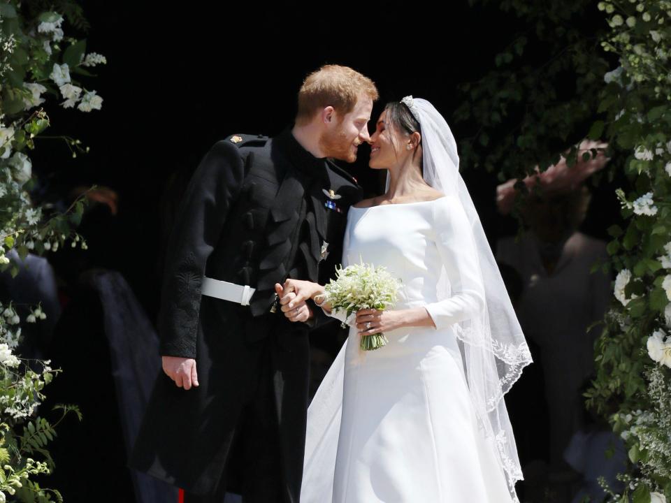 Harry in a black military uniform leaning in to kiss Meghan in a white wedding dress and veil standing outside surrounded by greenery.