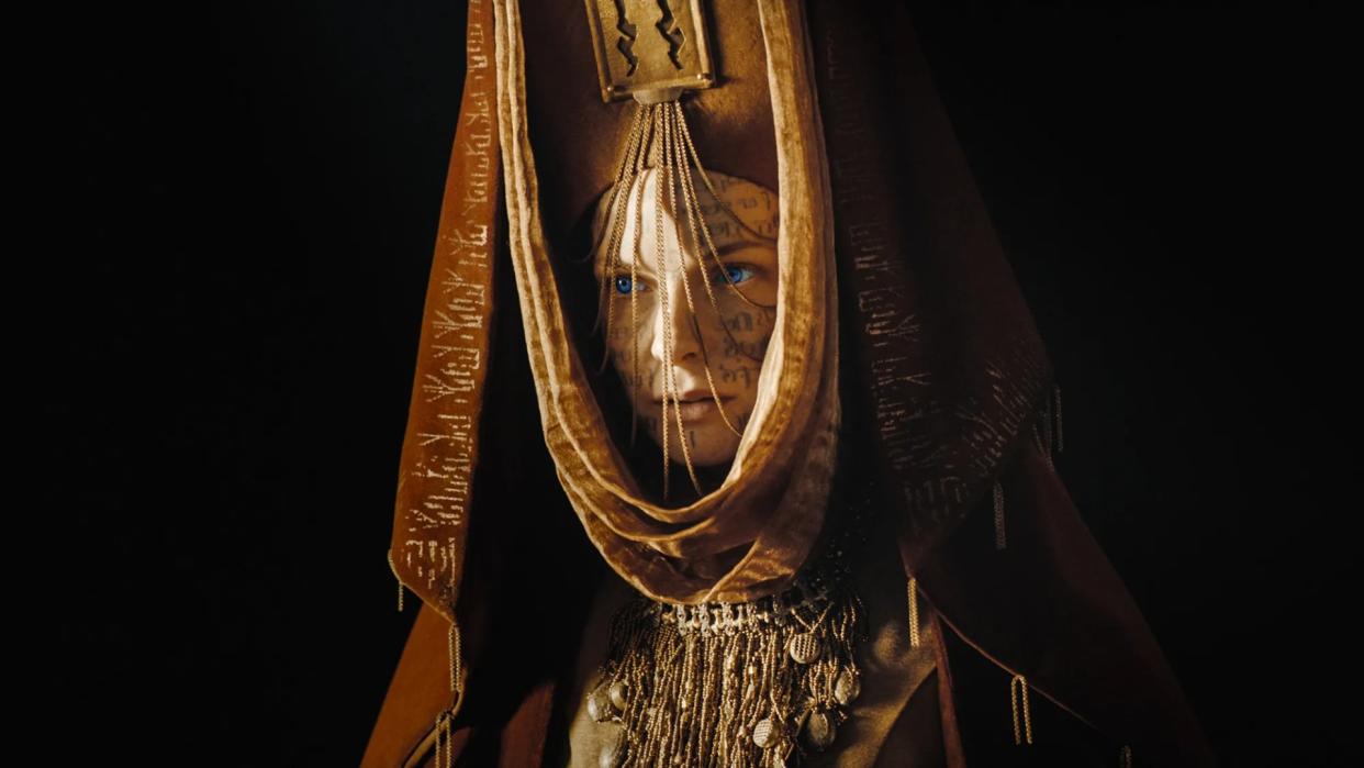  A woman with tattoos on her face wears an intricate crown with gold chains hanging down over her face. 