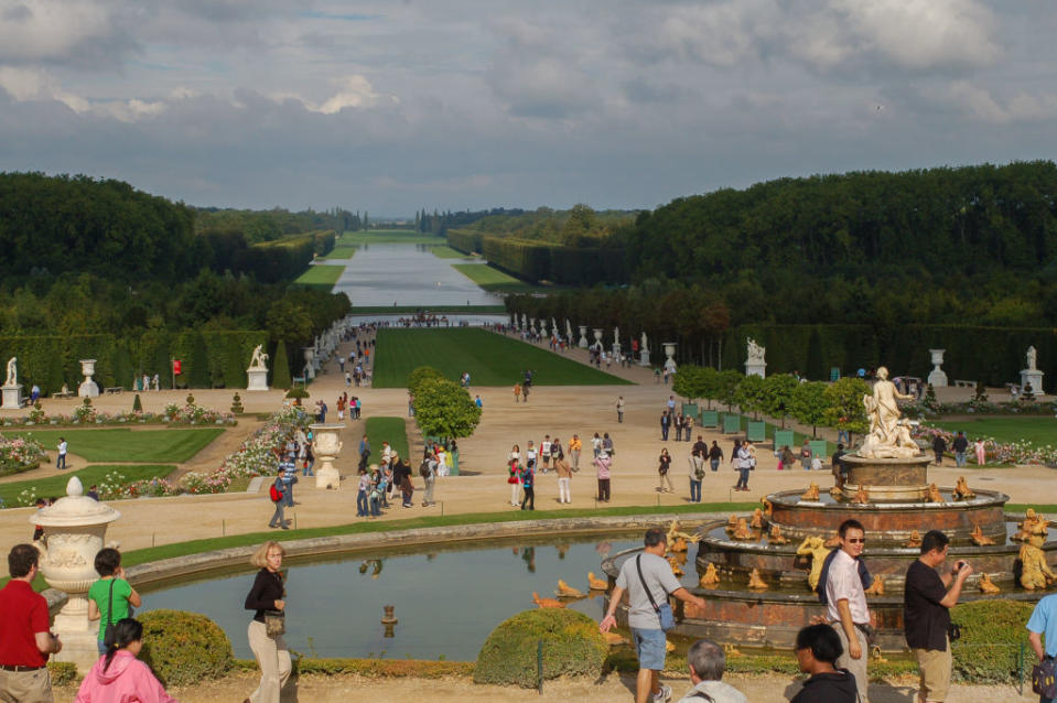 Crowds of tourists explore the vast gardens of the Palace of Versailles, with statues, a large fountain, well-maintained paths and wooded areas visible